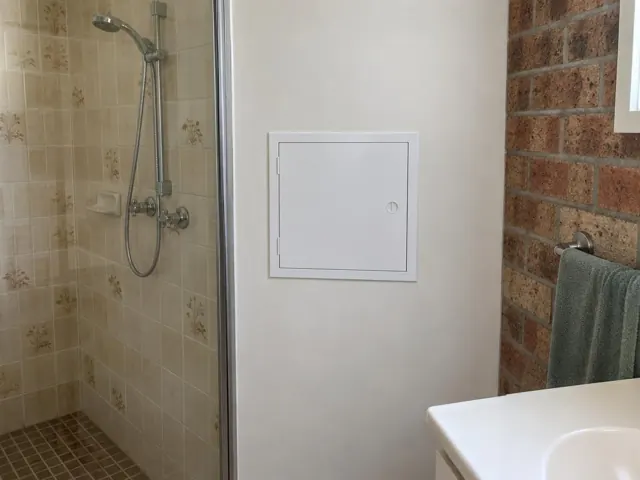 Small white access panel on a bathroom wall beside a shower and vanity, with a brick accent wall in Northmead